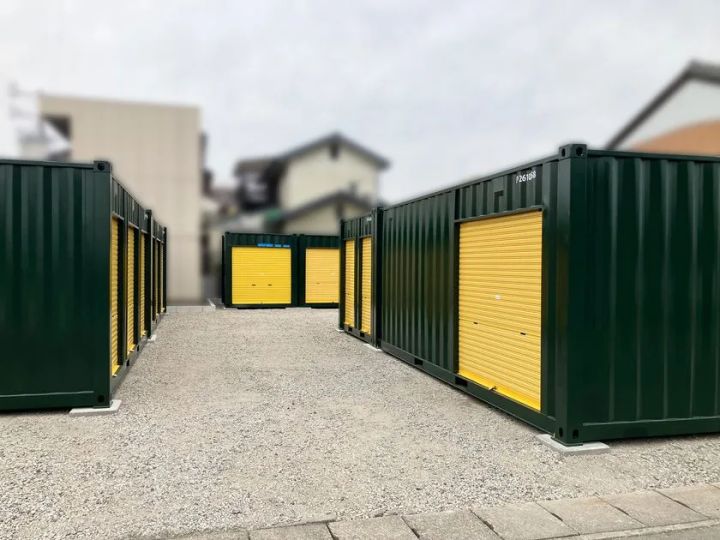 スペースプラス関弥生町の敷地内を通路中央から撮影した現場写真。左右に並ぶ緑色コンテナと黄色シャッターの個別ユニットが複数見え、砂利敷きの通路と住宅街の背景が確認できる。