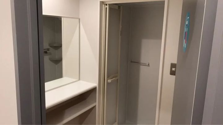 フィットネスジムの更衣室内洗面・着替えエリア。大型ミラーと棚付き洗面台、扉付きの個室着替えブース、壁面のシャワーサインが確認できる