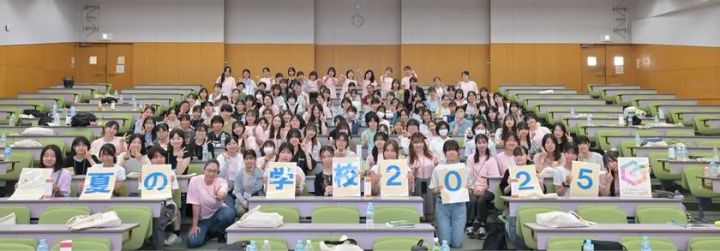 「夏の学校2025」の文字を掲げた女子中高生・スタッフ約100名が大学の講義室で集合した全体記念写真