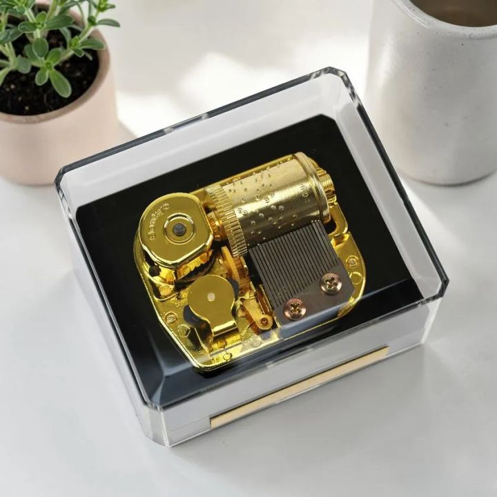 ゴールド製オルゴールムーブメントをクリアアクリルケースに収めた商品の俯瞰ショット。シリンダーと金属製コームが露出した内部機構が見える