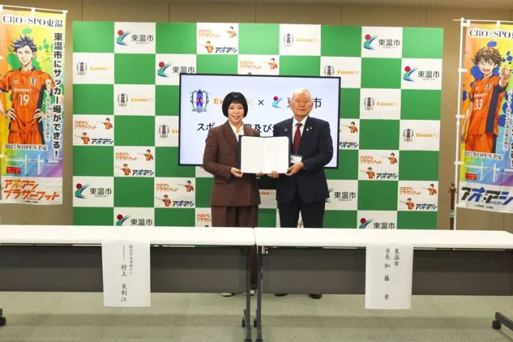 愛媛FC代表取締役社長・村上茉利江と東温市長・加藤章が協定書を手に並んで立つ調印式の2ショット写真。背景に「CRO×SPO東温」「アオアシ」のバナーと東温市・愛媛FCのロゴが並ぶ