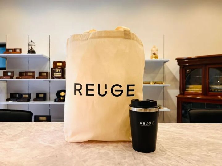 REUGEロゴ入りキャンバストートバッグと黒いステンレスタンブラーを店頭カウンターに並べたノベルティ2点セット。背景の棚にはREUGEオルゴール複数台が陳列されている。