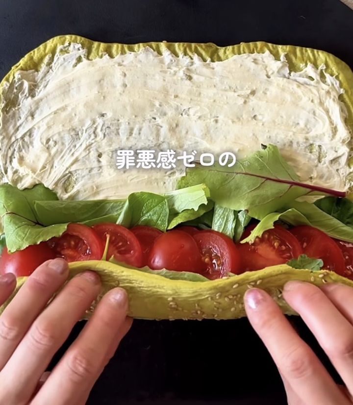 Wrap sandwich being assembled: yellow flatbread filled with cream cheese spread, lettuce, and sliced tomatoes, held open by hands.