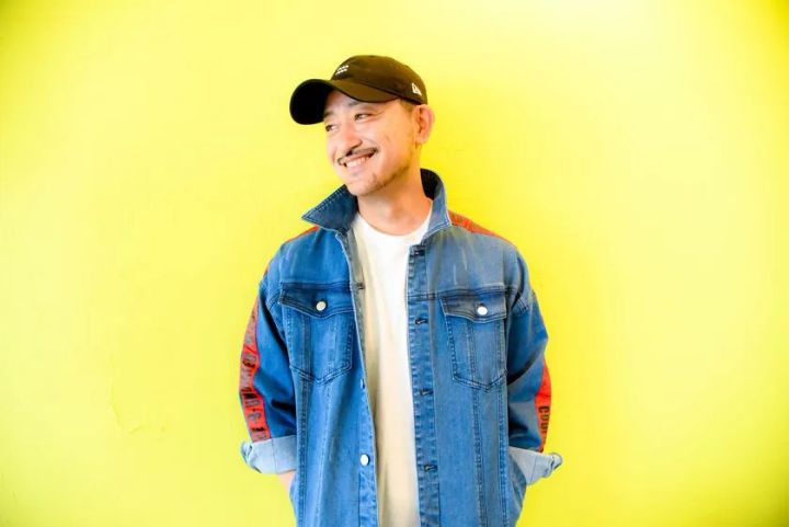 黄色背景の前に立つ男性のポートレート写真。デニムジャケットと黒キャップを着用し、笑顔でカメラ横を向いている宣伝・出演者系のプロフィール写真