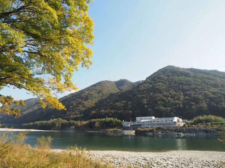 吉野川沿いの河川敷と山を背景に、株式会社オカベの白い工場建物を望む自然環境の外観ショット