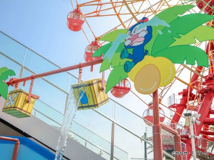 ヤシの木型オブジェにドキンちゃんが乗り、木桶から水が流れ落ちるびっくりバケツ仕掛けのアトラクション。背景に観覧車が見える屋外スペースでの撮影。