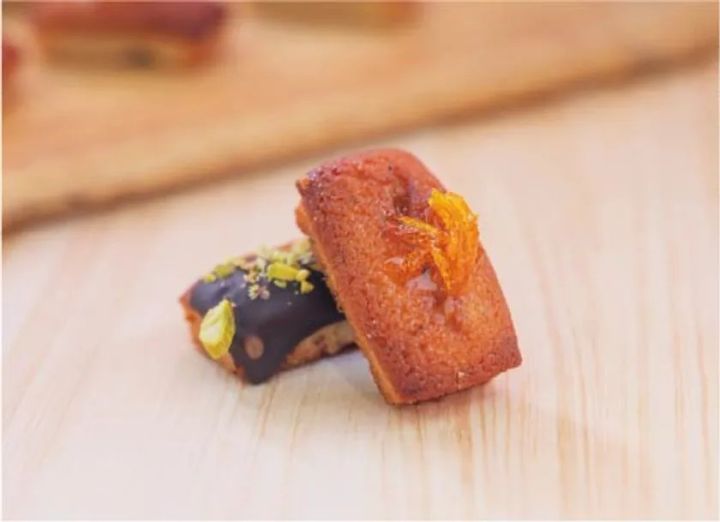 夏みかんの皮をトッピングした焼き色のフィナンシェ1個と、チョコレートコーティングにピスタチオをまぶしたフィナンシェ1個の計2種を木目台の上に重ねて配置した商品写真
