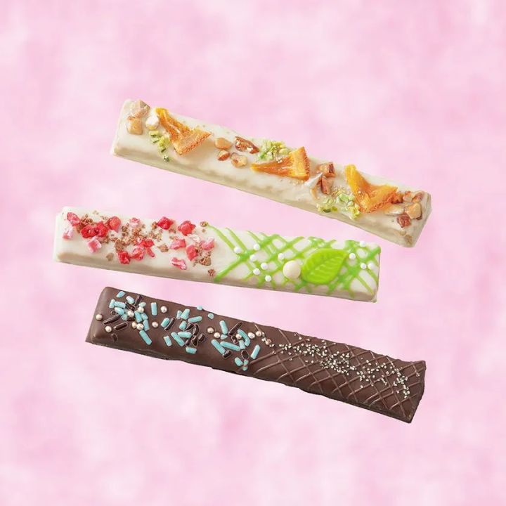 細長いチョコレートバー3種（ホワイト×ドライフルーツ・ミントグリーンデコ・ダーク×水色スプリンクル）をピンク背景に斜めに配置した3フレーバー比較ショット