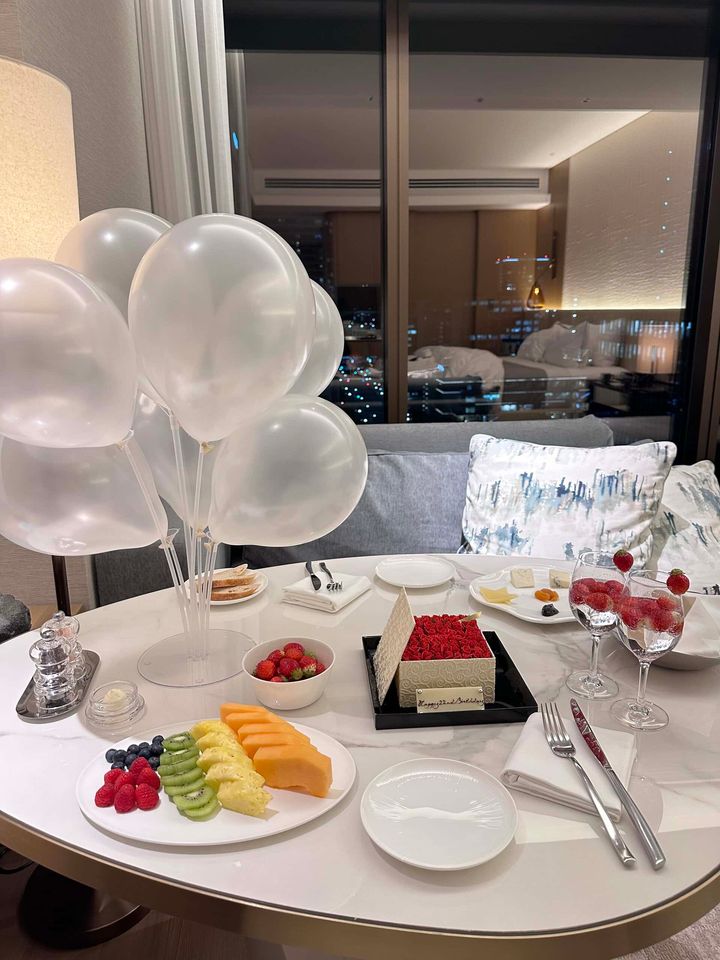クリアのバルーン、フルーツ盛り合わせ、ケーキ、ドリンクなどで装飾されたホテルの部屋のテーブル。大きな窓からは夜景が見える。