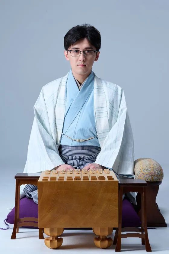 和服（白羽織・水色着物）姿の若い男性棋士が将棋盤の前に正座し、駒台と駒袋を両脇に置いたグレー背景のポートレート写真
