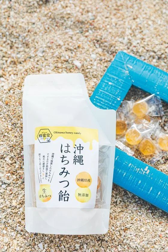 蜂蜜堂の沖縄はちみつ飴スタンドパウチ（沖縄県産・無添加・生はちみつ表記）と個包装キャンディが砂浜に置かれた商品写真