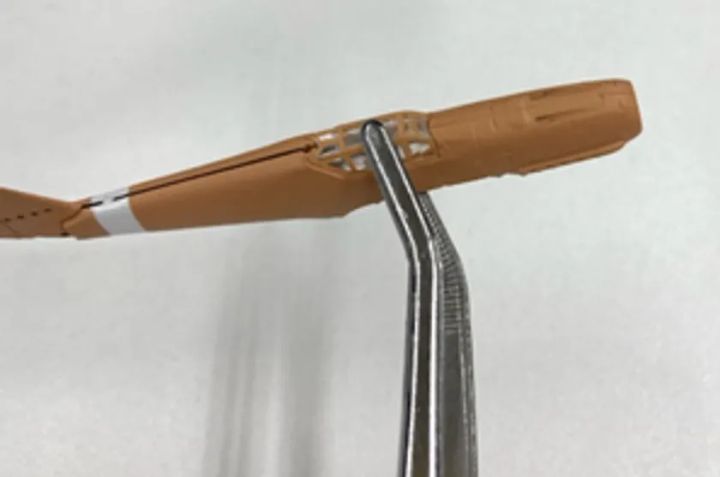 飛行機プラモデルの茶色い胴体パーツにステンレスワイヤーを差し込む工程を接写し、アロンアルフア光による金属と樹脂の接着デモを示すショット
