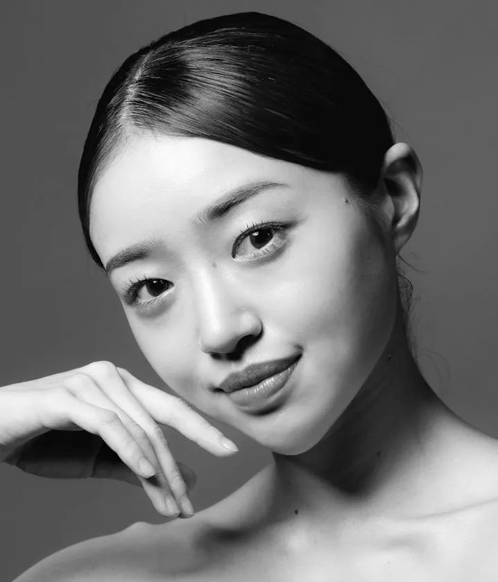 バレエダンサーと思われる女性のモノクロ正面ポートレート。髪をバレエバンにまとめ、頬に手を添えて柔らかい表情を見せる