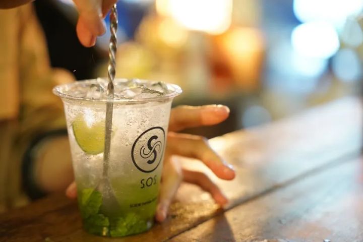 バーテンダーがSOSロゴ入りプラスチックカップに入ったライムとミントのモヒート系カクテルをマドラーでステアしているクローズアップショット