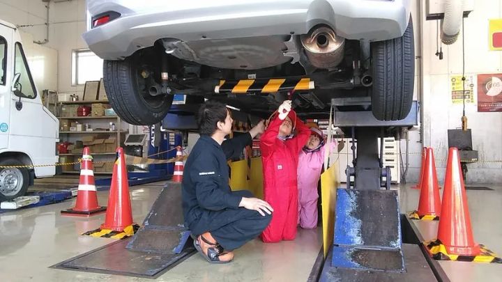 整備工場のリフトで持ち上げた車の下に入り、整備士と子ども2人がつなぎ姿で車両底部を懐中電灯で点検している様子