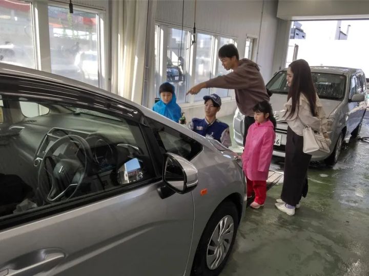 整備工場内で制服姿のスタッフが家族連れ（子ども2人・母親）に対して銀色の乗用車のボディを指差しながら説明している様子