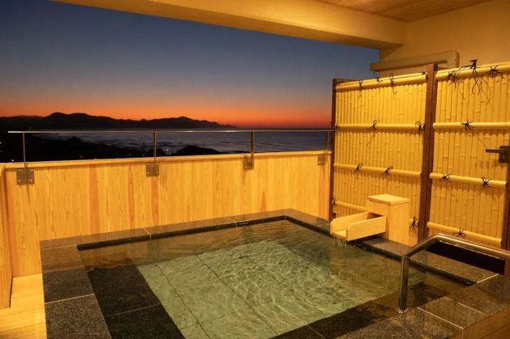 客室付き露天風呂（石造り・木製囲い）から夕焼けと日本海の水平線を望む夕景ショット。湯面と夕焼けのオレンジ色の空のコントラストが写る