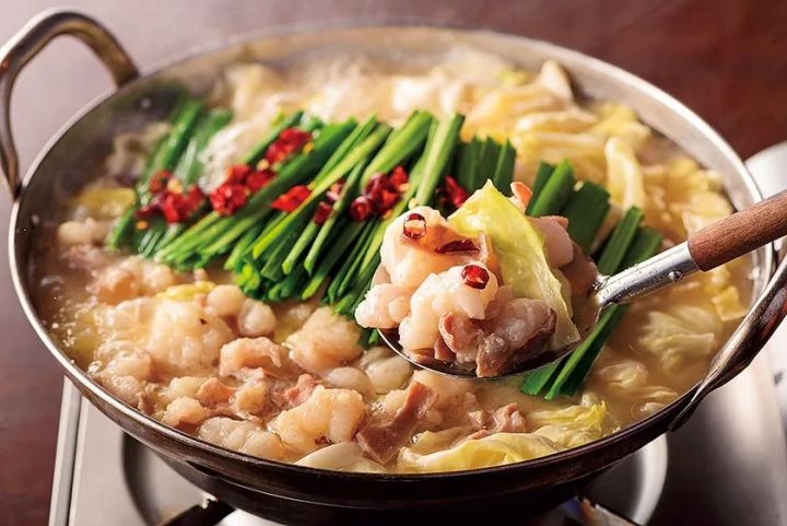 博多もつ鍋の鍋料理写真。牛もつ・白菜・ニラ・唐辛子が入ったスープを煮立たせた状態で、スプーンにもつを盛り上げた構図