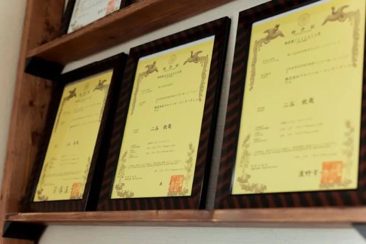額縁に入った特許証が木製棚に3枚以上並んで展示されている写真。二谷の名前が記載された黄色い特許証書が複数確認でき、技術的裏付けを示す。