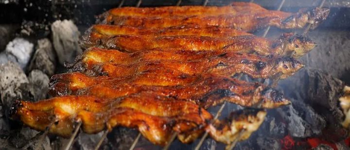 備長炭の炭火の上でタレを塗りながら地焼きされているうなぎの串が複数本並び、煙が立ち上る焼き場のクローズアップショット