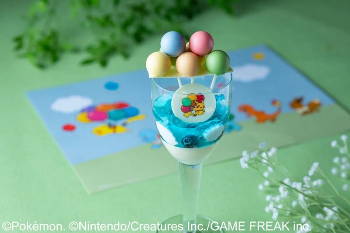 青いゼリーとクリームを重ねたグラスデザートの上部にパステル5色のケーキポップ球が串刺しで飾られ、ピカチュウとバルーン柄のチョコレートメダルが側面に添えられたポケモン30周年スイーツ