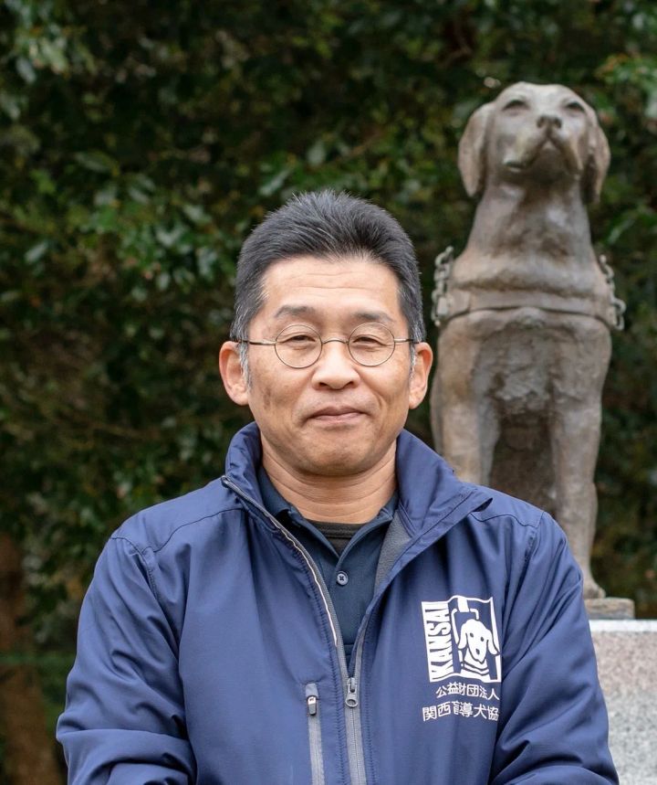 公益財団法人関西盲導犬協会のジャケットを着た男性が、盲導犬の銅像を背景に正面を向いて立っているポートレート写真