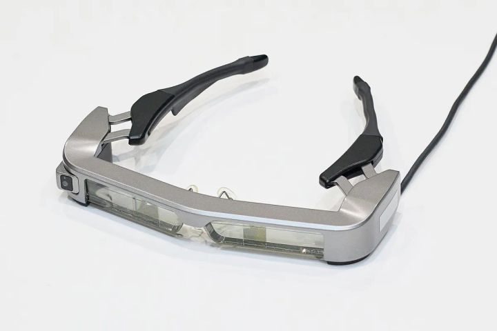 視覚障がい者向け道案内デバイスのスマートグラス本体。シルバーフレームにレンズユニットとカメラを内蔵し、黒いテンプル（つる）とケーブルが接続された斜め俯瞰の製品単体ショット。