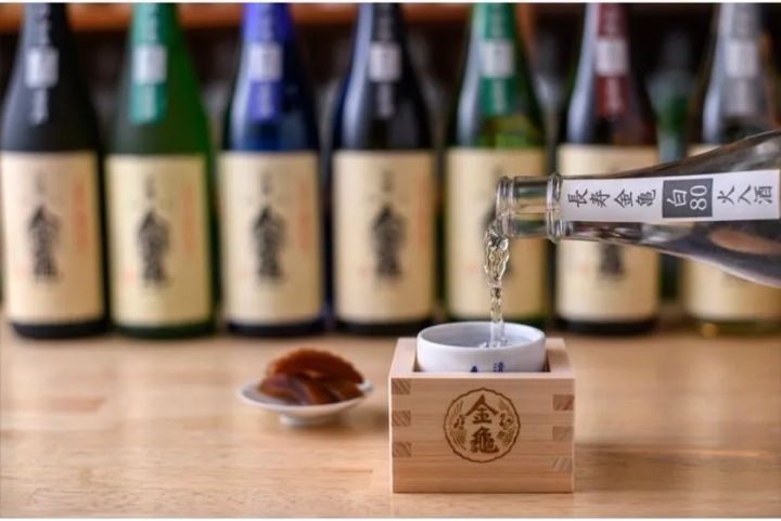 金亀の升と猪口に日本酒を注ぐ試飲シーン。背後に金亀ラベルの瓶が複数本並び、酒蔵見学・試飲体験の雰囲気を伝える写真
