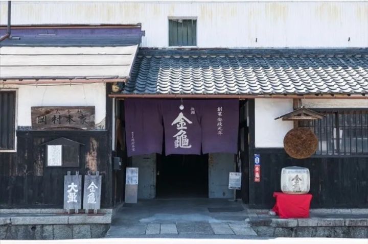 創業安政元年の酒蔵「金亀」（岡村本家）の外観写真。紺地に金亀と書かれた暖簾、鏡餅形の酒樽、瓦屋根の白壁蔵が写る