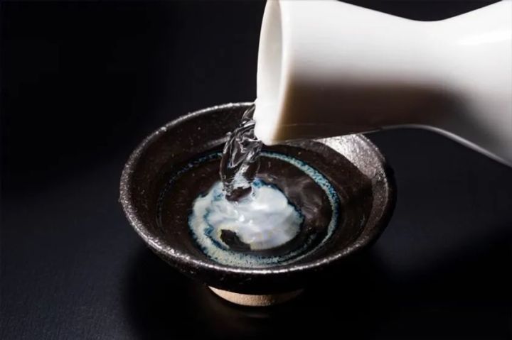 白い徳利から黒釉の盃に日本酒を注ぐクローズアップ写真。黒背景に液体の流れが際立ち、清酒の透明感と注酒の瞬間を捉えた演出カット