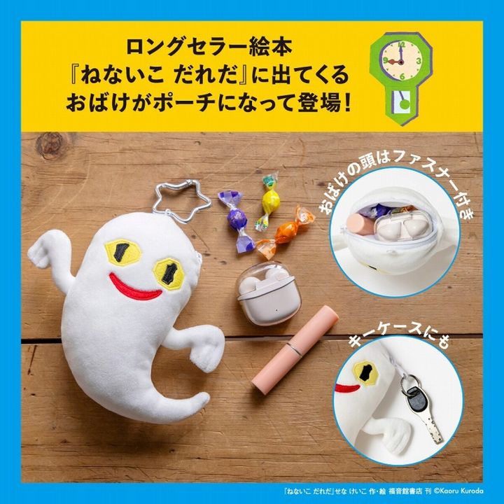 木目の床の上で、星形カラビナを付けた黄色い目と赤い口の白いおばけぬいぐるみポーチが横たわり、隣に包み紙キャンディとAirPodsケース、ピンクのリップが並ぶ俯瞰写真