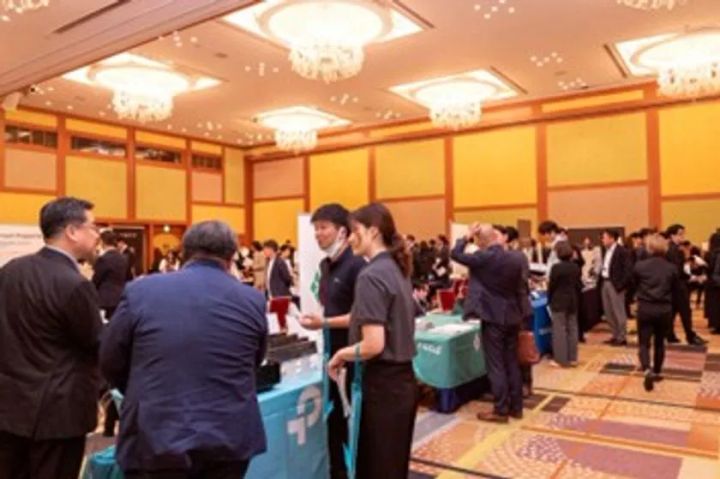 ホテル業界向け展示会の出展ブースエリアの実写。複数のブーススタッフが来場者と対面で商談・説明を行う様子が映り、ホワイトパネルのブース什器や青色テーブルクロスが確認できる