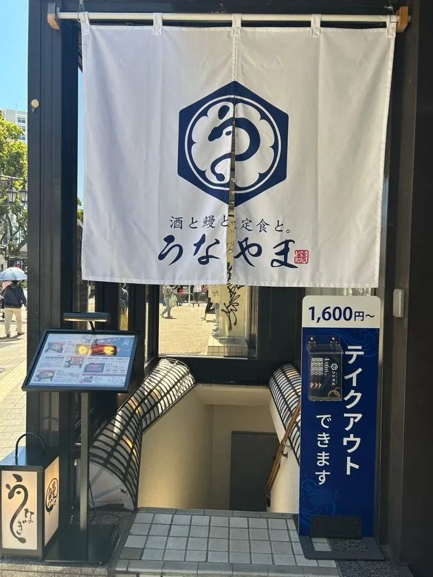 うなやまの店舗正面外観。白い暖簾に店名ロゴと酒と鰻と定食との文字、1600円〜のテイクアウト案内看板とメニューボードが確認できる