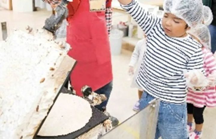 ヘアネットを着けた子供2人がスタッフと一緒に工場内でパン生地の成形体験をしている様子。コンベア上に伸ばされた生地が確認できる