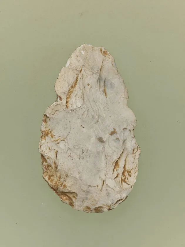 旧石器時代の握斧（ハンドアックス）または礫器を緑灰色背景で単体撮影。白色チャート質の扁平な楕円形石器で、表面に複数の剥離痕と錆色の鉄分変色が見られる