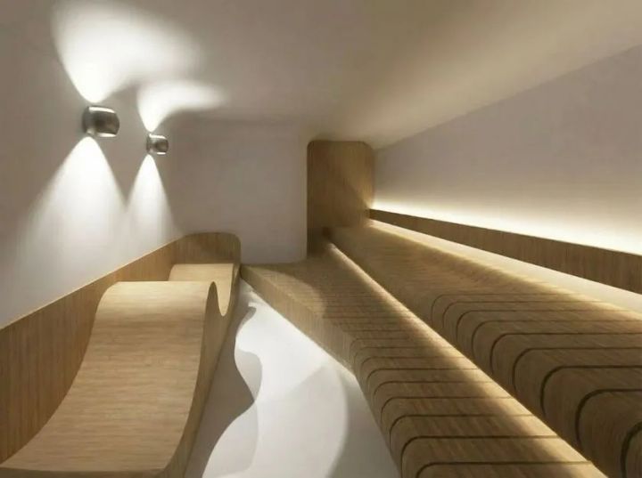洞サウナ(DO SAUNA)室内の木製段状ベンチと間接照明を正面から撮影した施設内観写真。曲線形状の木製ベンチ2列と壁面ウォールライト2灯が確認できる