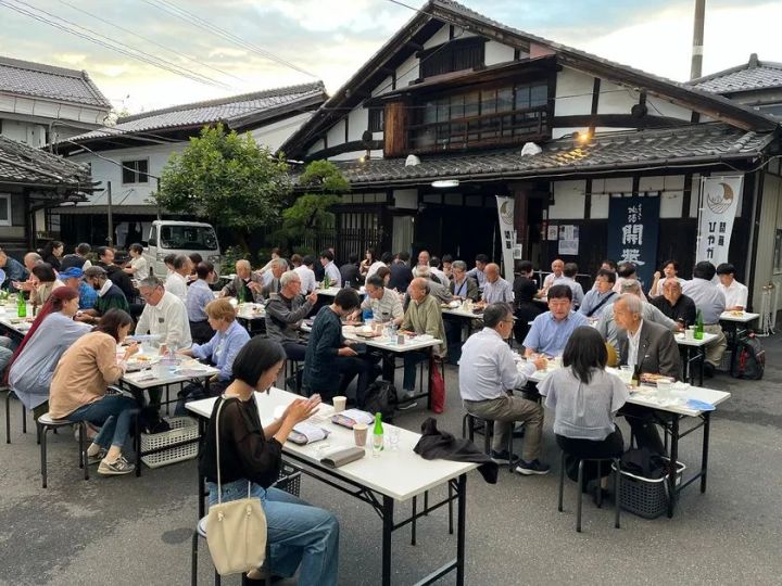 第一酒造敷地内で昼夕間に開催されたひやガーデンの屋外飲食風景。古民家風の酒蔵建物を背景に、参加者多数がテーブルに着席して食事と日本酒を楽しんでいる様子
