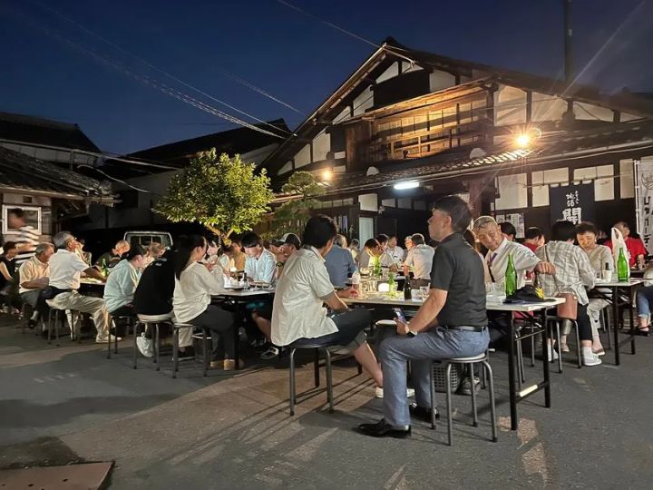 夜間のひやガーデン開催時の屋外宴席風景。ライトアップされた酒蔵前で参加者が日本酒瓶を囲んでテーブルごとに着席し、夜の酒蔵ガーデンでの飲食シーンを写している