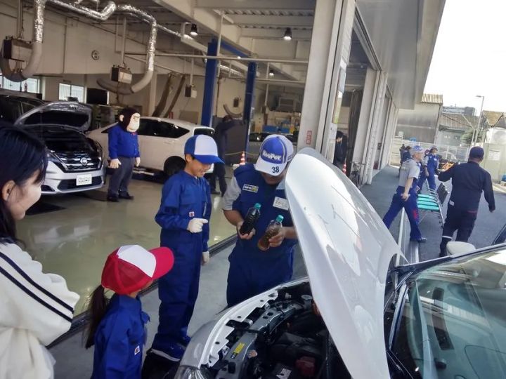KeePer技研のキッズ整備士体験イベントで、青い作業着と帽子を着た子どもたちがスタッフからエンジンオイルの確認方法を教わっているエンジンルーム前の場面
