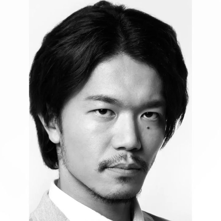 口髭とスタブルを持つ日本人男性のモノクロポートレート。白背景でスーツ襟が見え、強い眼差しが特徴的な演出家または出演者のプロフィール写真