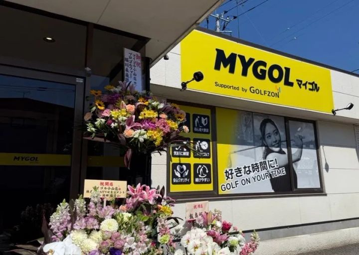 マイゴル小山店の店舗正面外観。黄色の「MYGOL」看板とGOLFZONロゴ、祝開店の花束、「好きな時間に！GOLF ON YOUR TIME」のキャッチコピーが掲示されたガラス面が写る