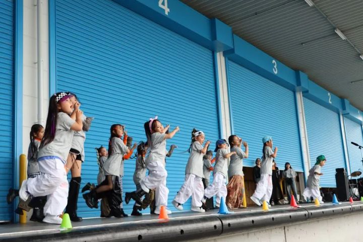 篠原陸運トラックステーションの青いシャッター前のステージで子どもたちがグレーと白のヒップホップ衣装でダンスパフォーマンスを披露している様子
