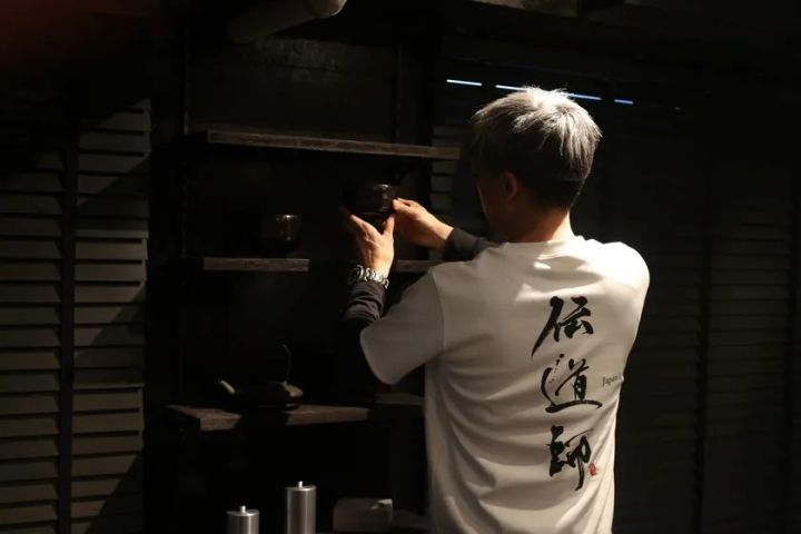 背中に「伝道師」の毛筆文字が入った白Tシャツを着た男性が、暗い和風の棚に黒い茶碗を配置している後ろ姿のスタッフ撮影カット