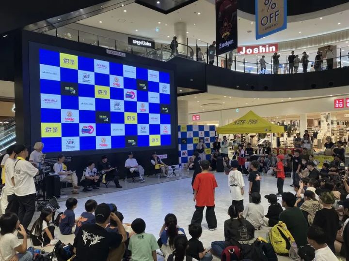 イオンモール館内の吹き抜けコートで開催されたブレイキン大会の競技風景。参加選手2名がフロアで向き合い、審査員席・大型スポンサーLEDビジョン・多数の観客が写る