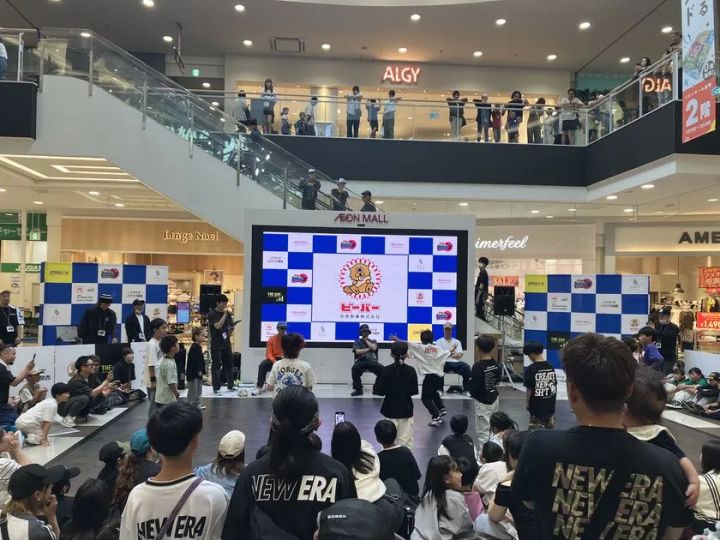 イオンモール館内コートでのブレイキン大会で複数選手がフロアに集まりパフォーマンス中の場面。スポンサーロゴ入りLEDビジョンと2階ギャラリーを含む多数の観衆が囲む