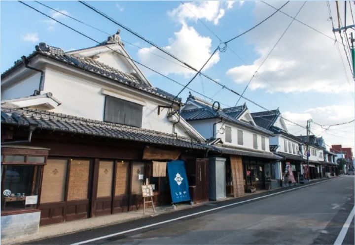 福岡・八女の伝統的な商家の町並みを撮影した外観写真。瓦屋根の白壁蔵造り建物が通り沿いに連続し、藍色の暖簾を掛けた店舗が展示会場周辺の雰囲気を伝える