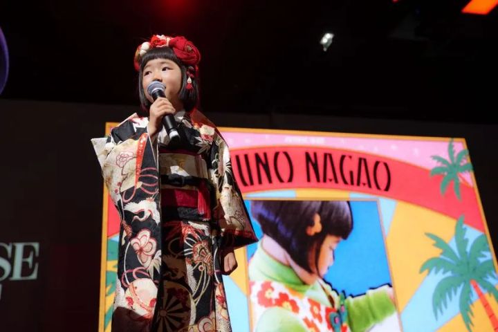 黒地に花柄の振袖と赤い髪飾りを着けた少女がマイクを持ちステージで歌唱し、背後にUNO NAGAOの名前と本人のカラフルなポスタービジュアルが映し出されている