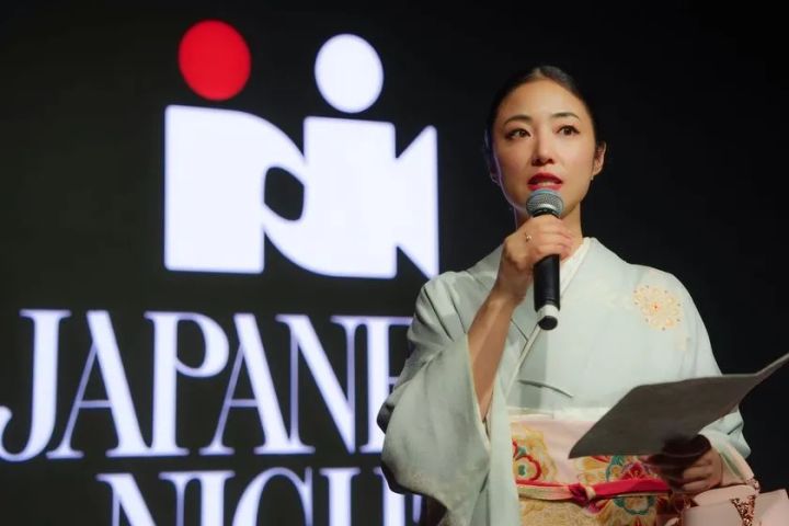 着物姿の女性がマイクを持ち手元の用紙を確認しながらスピーチするステージ上のショット。背景にJAPANESE NIGHTのロゴが大きく表示されている