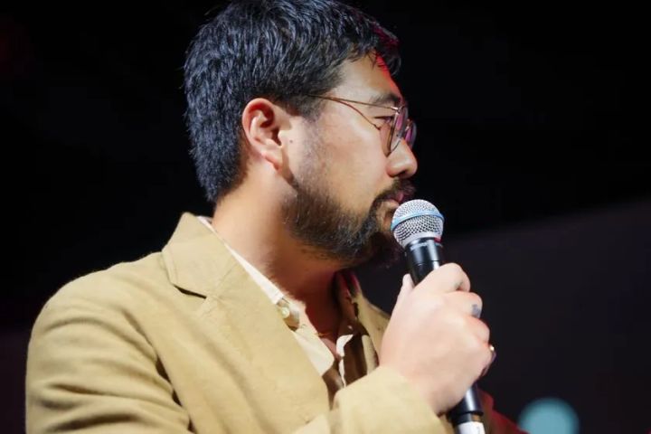 茶色いジャケットにメガネをかけた男性がマイクを口元に近づけステージ上で発言しており、暗転した背景にスポットライトが当たっているイベント登壇シーン
