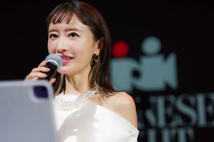白のオフショルダードレスにパールネックレスを着けた女性がマイクを手にステージで司会または挨拶を行い、背後にJAPANESE NIGHTのロゴが確認できる